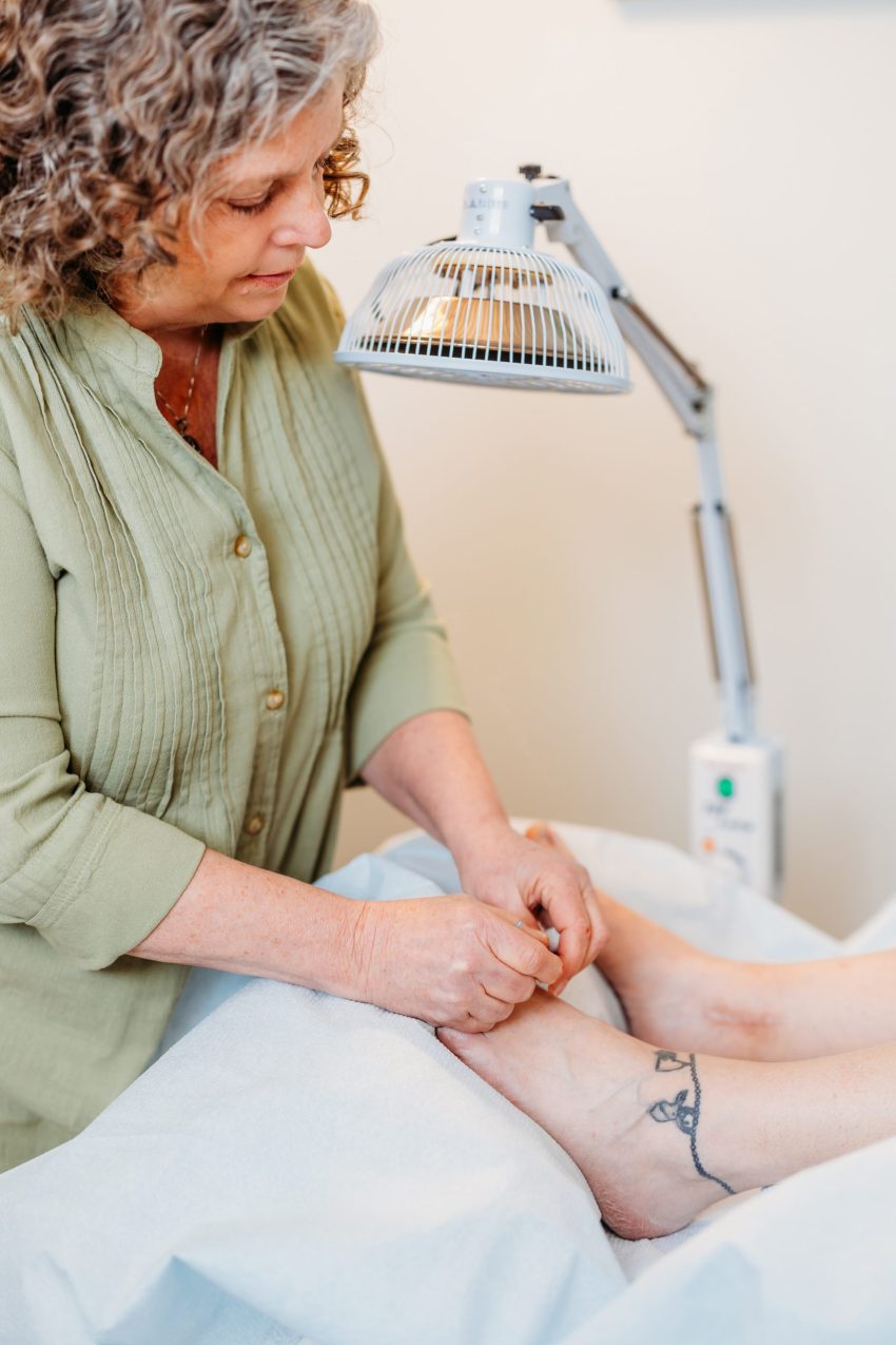 Maggie shot 5 Maggie Barili inserting needles into a patient’s foot during a treatment session, with a heat lamp overhead and calming clinical setting.