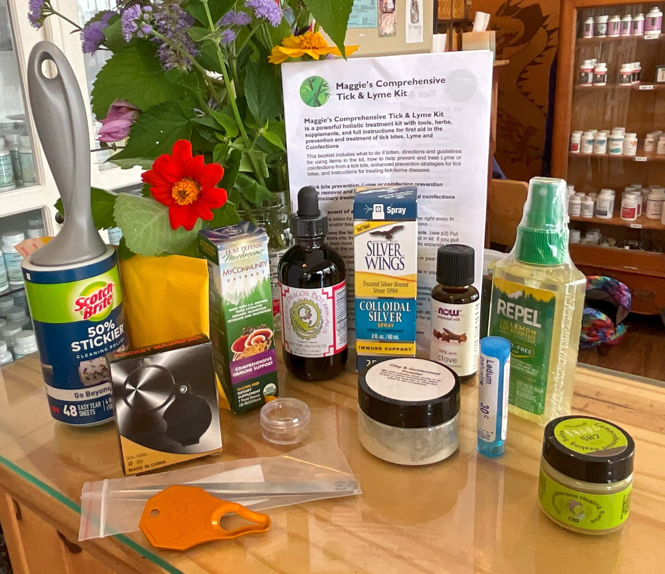Display of Maggie’s Comprehensive Tick and Lyme Kit, featuring natural remedies, herbal tinctures, repellents, homeopathics, and a detailed instruction sheet on a counter with fresh flowers.
